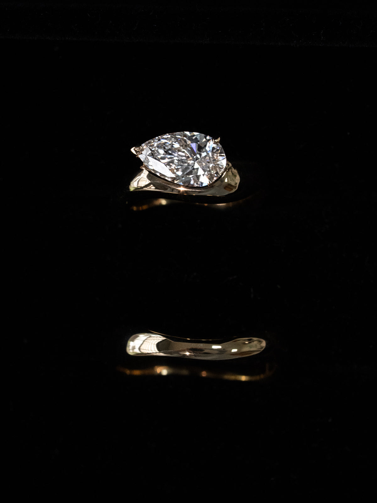 Overhead view of a custom sculptural engagement ring set with matching wedding band in 18k yellow gold, featuring a pear-cut diamond on the engagement ring, displayed on a light grey surface highlighting the flowing, curved design of both pieces.