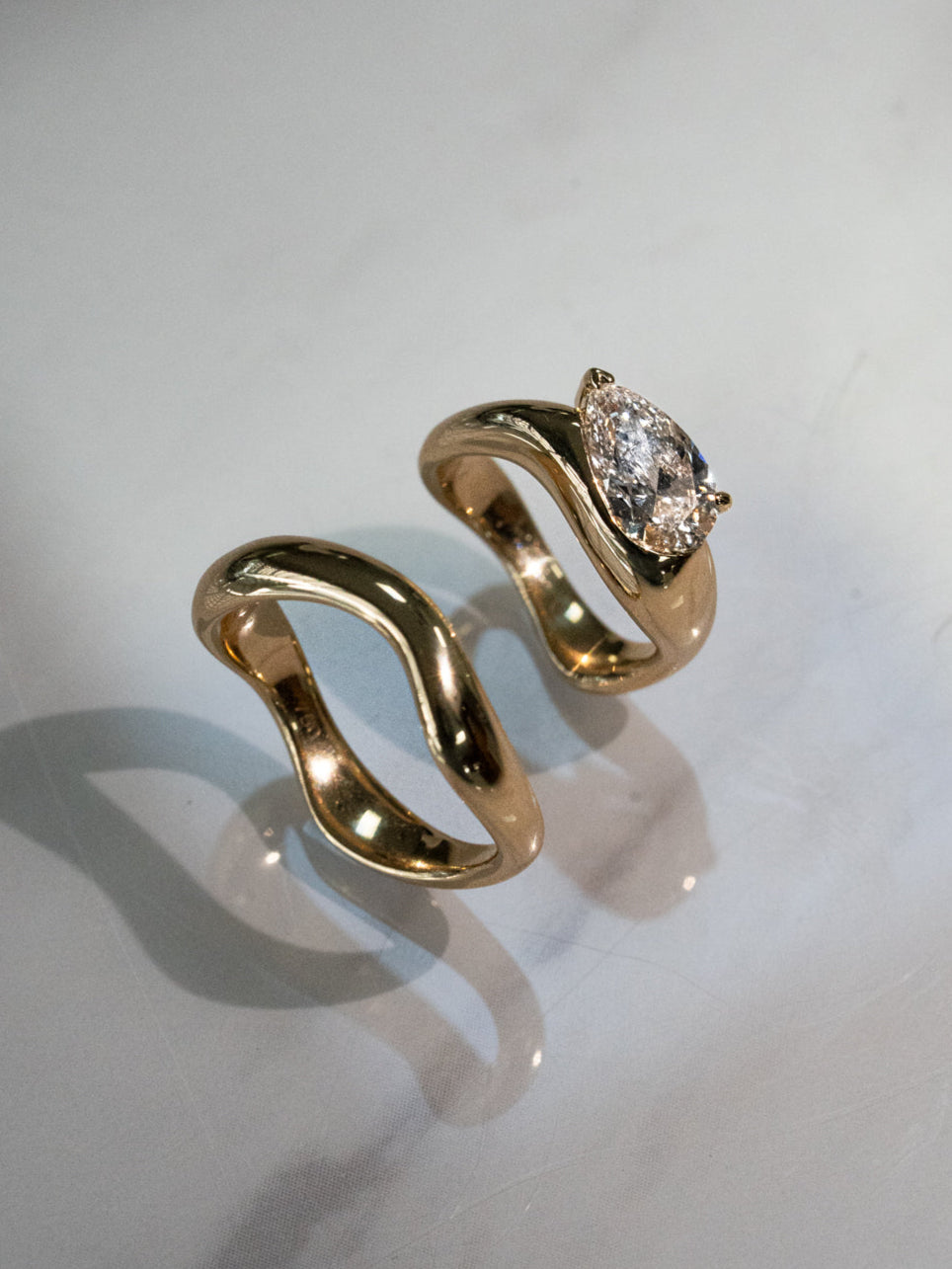 Overhead view of a custom sculptural engagement ring set with matching wedding band in 18k yellow gold, featuring a pear-cut diamond on the engagement ring, displayed on a light grey surface highlighting the flowing, curved design of both pieces.
